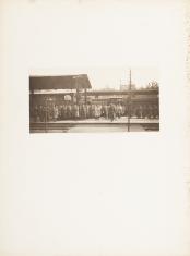 Fotografia wykonana w kolorystyce sepii, poziomy kadr. Grupa mężczyzn w długich płaszczach stojących na peronie dworca kolejowego. Po lewej stronie kadru widnieje daszek wiaty peronowej. W tle dalsza część zabudowy dworca oraz jasne niebo.