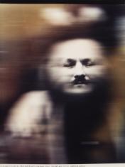 Kadr pionowy. Fotografia o dominującym odcieniu sepii. Portret mężczyzny, popiersie. Wyraźna jest jedynie środkowa część twarzy od czoła po brodę. Pozostała część zdjęcia jest rozmyta, niewyraźna.