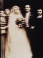 Utrzymana w kolorze sepii fotografia przedstawiająca stojącą w centrum parę w strojach ślubnych i troje gości za nimi, kontury są lekko rozmyte. 