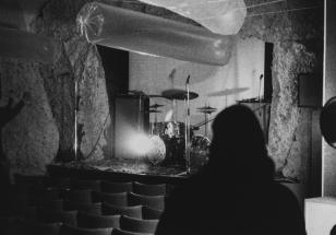 Czarno-biała fotografia ukazująca pustą scenę na której mikrofony i instrumenty perkusyjne. Nad sceną podwieszone obiekty o kształcie walców. Na pierwszym planie czarny zarys postaci z długimi włosami.
