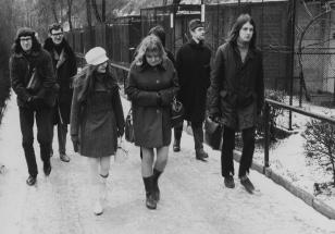 Czarno-biała fotografia ukazująca grupę osób w zimowych strojach idących po ośnieżonym chodniku w kierunku widza.