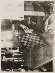 Fotografia wykonana techniką montażu, w centrum stół z szachownicą, przy nim mężczyzna i kobieta z głową wspartą na rękach, w dole po lewej fragment położony poziomo przedstawiający dwie postaci męskie we wnętrzu.
