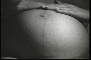 W czarno-białym kadrze widać nagi, napięty brzuch leżącej kobiety w zaawansowanej ciąży.