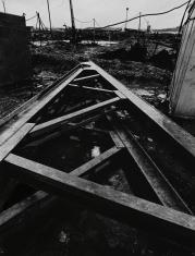 Fotografia czarno-biała, kadr pionowy. Fragment budowy. Na pierwszym planie fragment stalowej konstrukcji, w tle wykopy, słupy z rozwieszonymi przewodami.