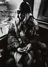 Fotografia czarno-biała, kompozycja w pionie. Kobieta o blond włosach, siedząca w rogu wewnątrz autobusu. Odziana jest w futro z lisa oraz ochronny kask na głowie. Wzrok skierowany ma w lewą stronę. Szyby autobusu mokre od zewnątrz.