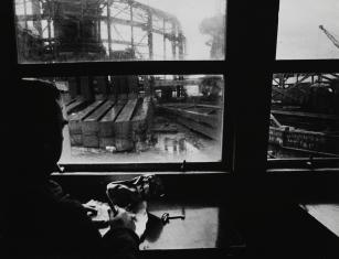 Fotografia czarno-biała, kadr poziomy. Na pierwszym planie przy lewej krawędzi, ciemna sylwetka mężczyzny zwróconego do widza plecami. Siedzi on przed oknem, przez które widać fragment budowy z metalowym szkieletem budynku.