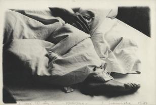 Fotografia czarno-biała, o orientacji pionowej. Fragment łóżka,  na którym leży przykryta pościelą kobieta. Spod pościeli widoczna jest jedynie stopa i dłoń. Fałdy tkaniny układają się w ostre zagięcia. Światłocień kontrastowy.