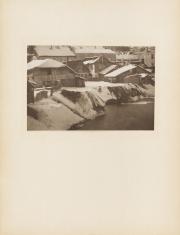 Fotografia wykonana w kolorystyce sepii, poziomy kadr. Widok na domy ze spadzistymi dachami, na których leży gruba warstwa śniegu. W prawej dolnej części obrazu, rzeka lub staw z brzegiem pokrytym śniegiem i miejscami zamarzniętą wodą.