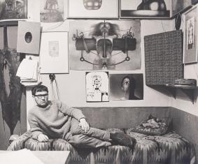 Fotografia czarno-biała przedstawiają Beksińskiego leżącego na łóżku w mieszkaniu, na ścianie zawieszone liczne rysunki, heliotypie i zdjęcie.