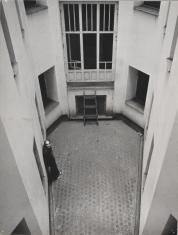Fotografia czarno-biała, ujęcie z góry na fragment wewnętrznego podwórka kamienicy o jasnym tynkowaniu wyłożony kostką brukową. Przy jednej ze ścian, po lewej stronie stoi mężczyzna oparty o nią.