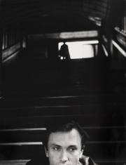 Pozioma fotografia czarno-biała. Na pierwszym planie w centralnej dolnej części twarz młodego mężczyzny ujęta od linii ust. Za nim	na drugim planie stoją ciemne schody z poręczami po obu stronach, a w tle jasna plama światła oraz zarys postaci.