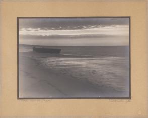 Fotografia czarno-biała naklejona bezpośrednio na ciemno-szary, zielonkawy papier i szarą tekturę, przestawiająca morze, fragment plaży i łódź.