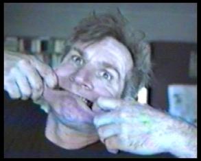 Kadr z filmu, przedstawiający wykrzywioną twarz artysty rozciągającego swoje usta z obu stron rękami.