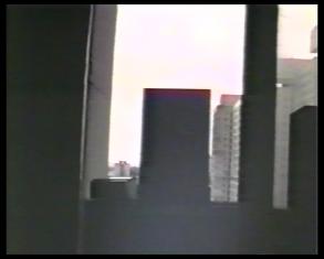 Kadr z filmu przedstawiający widok przez okno, za którym widać szare prostokąty wieżowców.