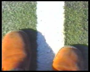 Kadr z filmu, przedstawiający spojrzenie z góry na czubki brązowych butów i asfalt ulicy z białą linią pionowo przecinającą kadr.