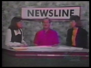 Fotografia przedstawia studio telewizyjne. Za szerokim pulpitem siedzą trzy osoby: dwie kobiety, w środku mężczyzna. Rozmawiają. Nad nimi znajduje się duża pozioma tablica z napisem drukowanymi literami Newsline.