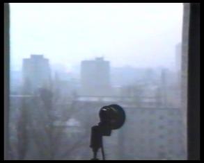 Kadr z filmu - przedstawiający ciemny kształt ciemnego przyrządu na tle okna, za którym widać osiedle szarych wieżowców.