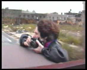 Kadr z filmu przedstawiający popiersie ciemnowłosego chłopca z aparatem fotograficznym, wychylonego z jadącego pojazdu, w tle rząd budynków.