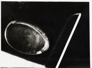 Fotografia przedstawia abstrakcyjną kompozycję, powstałą w wyniku zawieszenia w przestrzeni szklanego jajka i jasnego pręta ustawionego pionowo w stosunku do poziomej bryły jajko. Czarne tło.