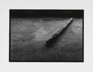 Fotografia czarno-biała, z prawej na podłodze z kwadratowych płyt leży drewniana belka, ukośnie w stosunku do krawędzi obrazu, całość ciemna.
