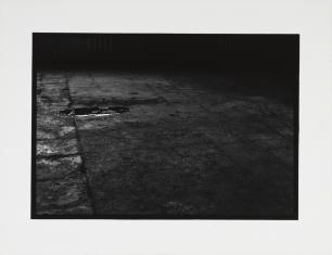 Fotografia czarno-biała, widziana z góry podłoga wykonana z kwadratowych, zniszczonych i popękanych płyt, z lewej powyżej środka rozlana plama ciemnej połyskującej cieczy, całość ciemna.