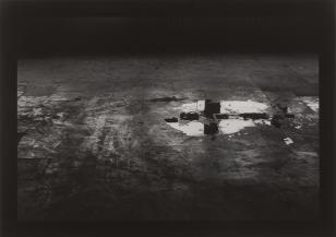 Fotografia czarno-biała, na widzianej z góry podłodze z lewej w środku duża kałuża, w której odbijają się okna.