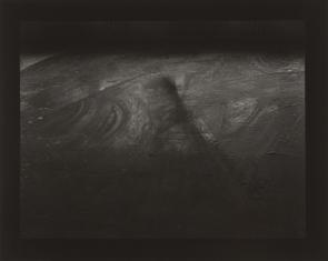 Fotografia czarno-biała, widziana z góry podłoga, widoczne ślady przesuwania czegoś długiego (belki) po niej, całość ciemna.