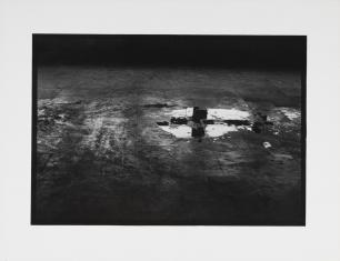 Fotografia czarno-biała, na ciemnej widzianej z góry podłodze z prawej rozlana kałuża, a w niej odbijające się fragmenty wnętrza, górna część obrazu ciemna, brak szczegółów w cieniach.