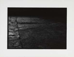 Fotografia czarno-biała, widziana z góry podłoga wykonana z kwadratowych, zniszczonych i popękanych płyt, całość ciemna.