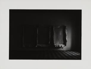 Fotografia czarno-biała, w ciemnym pomieszczeniu na ścianie wiszą trzy obiekty - naciągnięte luźno na blejtram płótno, z prawej światło padające z najprawdopodobniej okna, lewa strona ciemna.