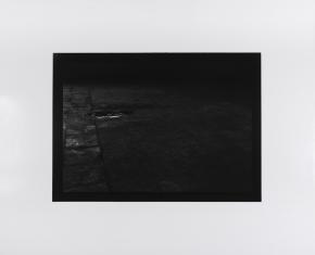 Fotografia czarno-biała, widziana z góry podłoga, lekko z lewej u góry ciemna mała kałuża, podłużna, reszta fotografii w cieniu.