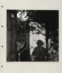 Fotografia plenerowa czarno-biała o formacie kwadratowym. Na drzwi drewniane w centrum obrazu pada cień postaci i liści z drzewa nad nią. Przy lewej krawędzi stoi drewniany płot z białą cyfrą 13. Między drzwiami, a płotem w tle ogród.