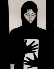 Fotografia przedstawia postać ubranej na czarno kobiety trzymającej przed sobą w czarnych rękawiczkach rulon białego papieru. Twarz jasno oświetlona.