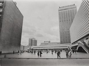 Fotografia czarno-biała, poziomy kadr z widokiem na Alexanderplatz w Berlinie. Zdjęcie budynków na tle zachmurzonego nieba. W tle rozpoznawalny wieżowiec: Stadt Berlin.