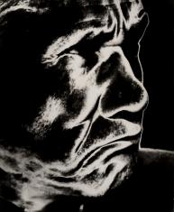 Fotografia przedstawia portret profilowy Zenona Kononowicza. Brwi silnie zmarszczone, niewidoczne w głębokim cieniu oczy, zaciśnięte i opuszczone w dół usta. Silne kontrasty czerni i bieli.