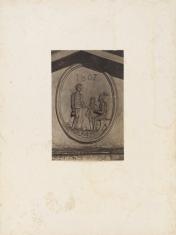 Fotografia wykonana w sepii, kadr pionowy. Zbliżenie na zdobienie budynku w postaci reliefu. Płaskorzeźba w elipsie przedstawia trzy osoby: kobietę, dziecko i siedzącego mężczyznę o kuli. Nad postaciami widnieje cyfra 1807.