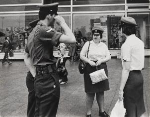 Fotografia czarno-biała, poziomy kadr, zdjęcie uliczne. Na pierwszym planie grupa policjantów rozmawiających na chodniku ( dwie kobiety w białych koszulach i dwóch mężczyzn w ciemnym umundurowaniu). W tle duża witryna sklepowa i przechodnie.