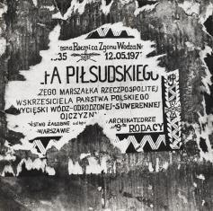 Fotografia czarno-biała, poziomy kadr, detal. Na ciemnej ścianie częściowo zdarta informacji o rocznicy śmierci marszałka Józefa Piłsudskiego. Na obrzeżach widać zdobienie z mundurów oficerskich Wojska Polskiego oraz order krzyża Virtuti Militari.
