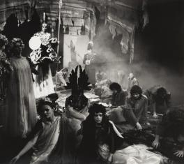 Fotografia czarno-biała, kadr zbliżony do kwadratu. Na pierwszym planie u dołu grupa siedzących postaci w różnych kostiumach o różnorodnym odcieniu. W tle kobieta jasno oświetlona trzymająca w lewej dłoni halabardę oraz tło przypominające budynek.