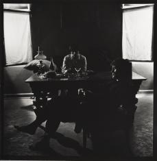 Fotografia czarno-biała, format bliski kwadratowi. W centrum dwóch mężczyzn siedzących przy ciemnym stole. Mężczyzna na pierwszym planie siedzi do widza lewym bokiem. Drugi za meblem. W tle przy górnych krawędziach dwa białe prostokąty. Centrum ciemne.