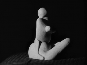 Czarno-biały kadr z filmu przedstawiający rzeźbę - jeden z aktów Katarzyny Kobro, odwroconą prawym profilem i wydobytą światłem z ciemnego tła.