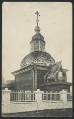 Czarno-biała fotografia przedstawia stojący za białym kamiennym płotem budynek drewnianej cerkwi zwieńczonej kopułą i wieżyczką.  To Zosimo-Zawwatiewski Chram w Zaczaczu.