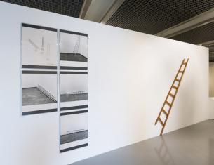 Instalacja konceptualna w przestrzeni sali wystawowej - po prawej stronie wisi na ścianie drewniana drabina w skrócie perspektywicznym, po lewej stronie pięć czarno-białych tablic - jedna projektowa i cztery zdjęcia dokumentacyjne.