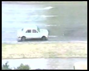 Kadr z filmu - widoczna z góry ulica z małym białym samochodem przy bocznej krawędzi wielopasmowej jezdni, poniżej pas trawnika i chodnika.
