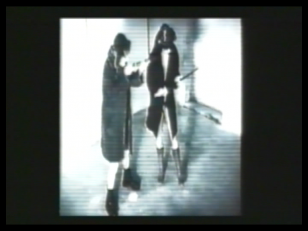 Kadr z filmu - czarnobiały, lekko rozmazany obraz dwóch postaci w ciemnych płaszczach na tle jasnych ścian.