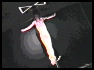 Zdjęcie zrobione z góry. Na podłodze leży krzyż. Rozpięty jest na nim nagi mężczyzna. Nad jego głową  wymalowane są dwa trójkąty stykające się wierzchołkami, przez co przybierają kształt przypominający nieskończoność.