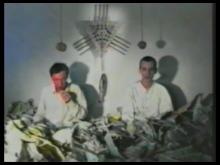 Kadr z filmu, przedstawiający dwie postacie w białych koszulach, za nimi wisi biały zgeometryzowany totem.