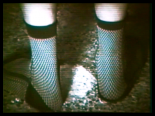 Kadr z filmu przedstawiający dwie nogi widoczne do połowy łydki, w skarpetkach w drobną kratkę z czarnymi ściągaczami, częściowo zanurzone w piasku.
