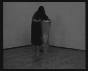 Kadr z czarno-białego filmu, przedstawiający ciemnowłosą kobietę w pomieszczeniu, schyloną nad wiadrem, trzymającą w rękach ścierkę.