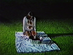 Kadr z filmu - w centrum kadru na tkaninie rozłożonej na trawniku klęczy ubrana na biało kobieta, przed nią znajduje się metalowa miska.